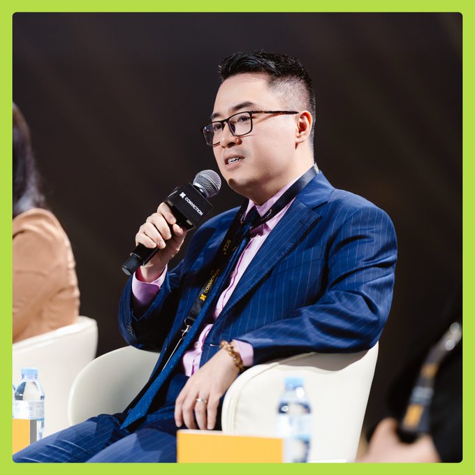 A bespectacled man in a blue pinstripe suit and pink shirt speaks into a "Conception"-branded microphone at Ho Chi Minh City Technology Day 2026. A lanyard around his neck displays "2025," consistent with his role as Deputy Head of Vietnam's Digital Asset Trading Market Management Board discussing investor regulations.