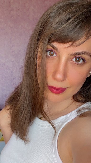 A selfie of a young woman with brown hair wearing red lipstick and a white tank top.