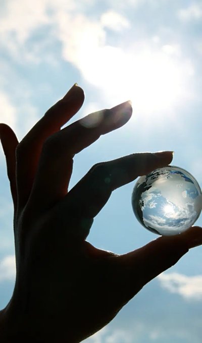 A silhouetted hand with long, manicured nails pinches a small crystal ball against a bright, cloudy sky.