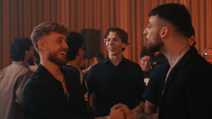 Two men warmly shake hands while smiling at each other during a social gathering, with other attendees mingling in the warmly lit background. The friendly, upbeat interaction aligns with the post's "GM" (good morning/good vibes) greeting.