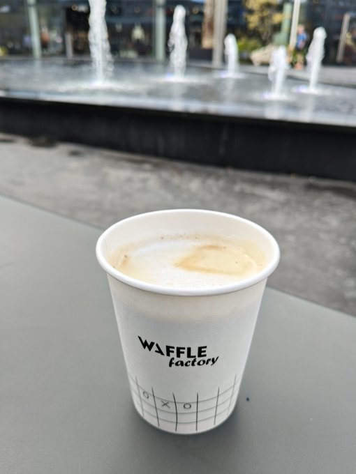 A paper cup from "WAFFLE factory" filled with a frothy latte, sitting on a gray table in front of an outdoor water fountain feature.