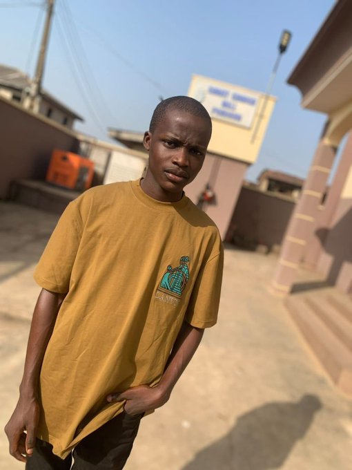 A young man in a yellow t-shirt stands outdoors, looking at the camera with a neutral expression.