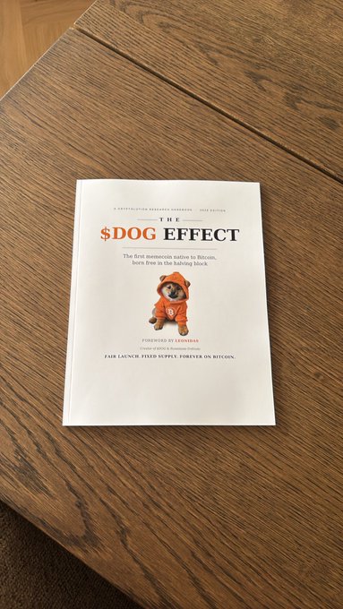 A book titled 'THE $DOG EFFECT' with the subtitle 'The first memecoin native to Bitcoin, born free in the halving block' rests on a wooden table, featuring a small dog in an orange hoodie on the cover.