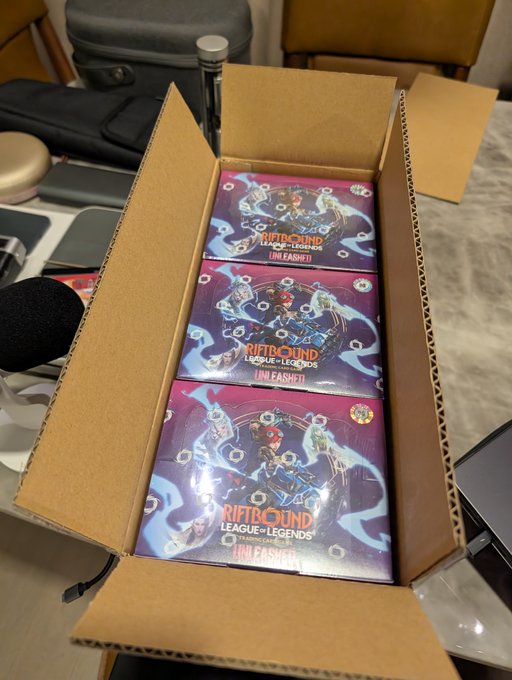 An open cardboard shipping box containing three sealed 'Riftbound: League of Legends Unleashed' trading card game booster packs.