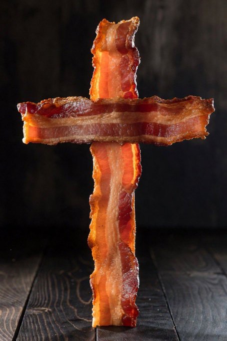 A crispy bacon cross stands upright against a dark wooden background, playing on the post's phrase "Stay strapped" by visually replacing a traditional ammunition cross belt with strips of cooked bacon.