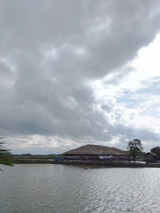 A cloudy sky hangs over a building with a thatched roof situated on the edge of a body of water.