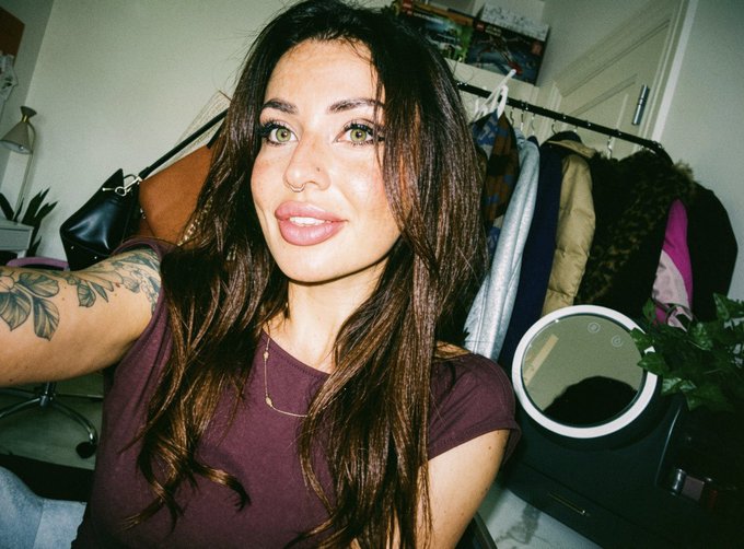Portrait of a woman with long dark brown hair wearing a maroon shirt, with a floral tattoo visible on her upper arm. She is in a room with a clothing rack and a round mirror in the background.