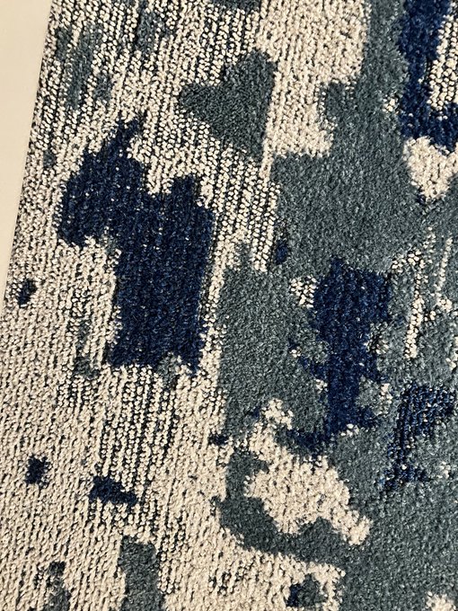 A close-up photograph shows the textured surface of a carpet or rug with an abstract pattern in shades of blue, navy, and white.