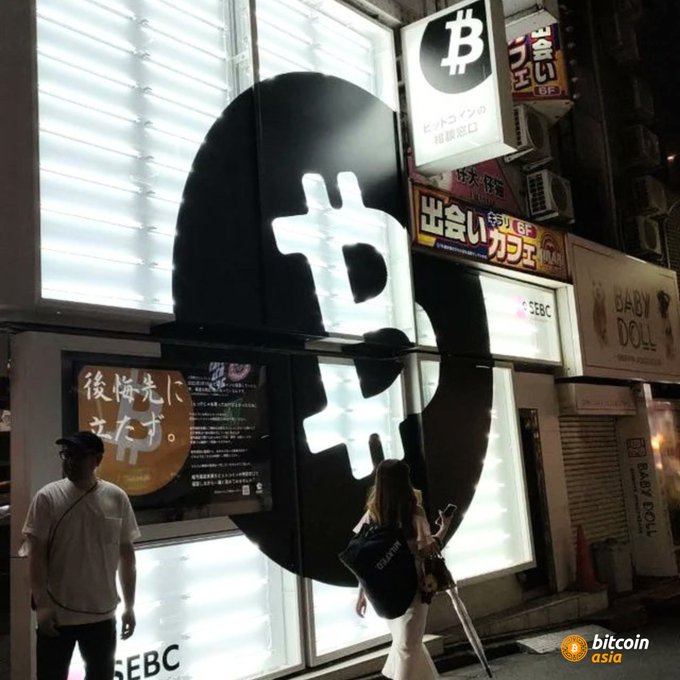 The image shows a large, illuminated Bitcoin sign displayed prominently on a building's facade. A man and woman are standing next to this sign, with one person holding an umbrella. The scene also includes other buildings and people walking by, indicating that they are in a busy urban area.