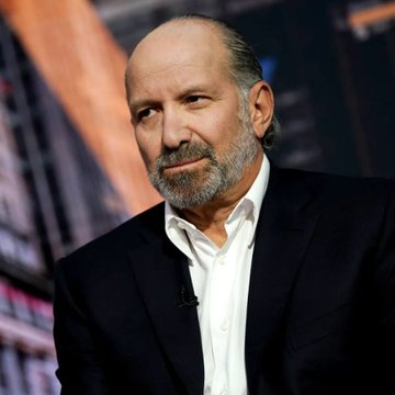 An older bearded man with a bald head wearing a black suit jacket and white shirt, appearing on what looks like a television panel.
