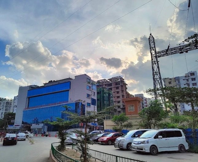An urban street scene showing a blue glass commercial building, parked cars, power line towers, and a dramatic cloudy sky.