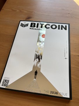 The image shows a white book with black text titled "BITCOIN Magazine" lying flat on a wooden table. The cover design includes an illustration of a man walking down a hallway or tunnel, which adds visual interest to the book's appearance. The book appears to be new and has a shiny finish, indicating it is in good condition.