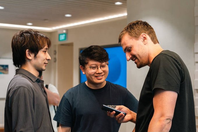 A photograph of three young men in casual attire interacting and smiling while one holds a smartphone, appearing to be at a tech meetup or office setting.