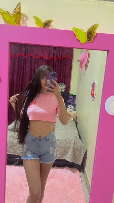 A mirror selfie of a young woman with long dark hair wearing a pink crop top and denim shorts, taken in a pink-decorated room featuring butterfly ornaments on the mirror and plush toys in the background.