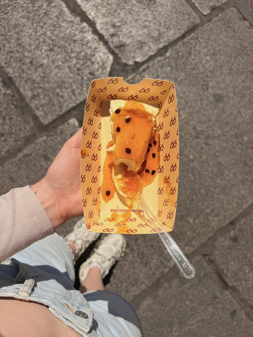 A first-person view of someone holding a paper tray containing a dessert with orange sauce and black seeds, eaten with a clear plastic spoon over a stone-paved ground.