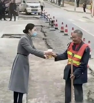 A woman wearing a face mask and grey coat hands a cup of coffee to a smiling elderly street sweeper in a high-visibility orange vest on a roadside.