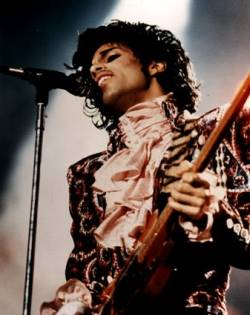 A vintage photo of a musician with long dark hair and a mustache, wearing a ruffled shirt and playing a guitar in front of a microphone.