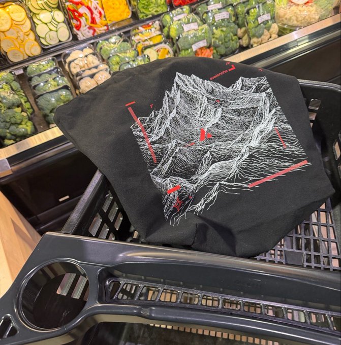 A black tote bag featuring a white and red abstract design rests in a shopping cart in front of a produce section stocked with vegetables like zucchini and broccoli.
