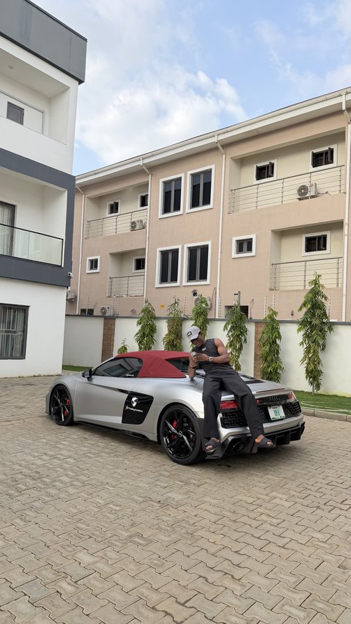 A man leans against a silver Audi R8 Spyder with a red soft top, parked on a paved driveway outside a beige residential building, as he checks his phone. His luxury ride serves as a visual flex to back his bold ₦20 million wager on crypto over forex, reinforcing the post's theme of high-stakes confidence and success.