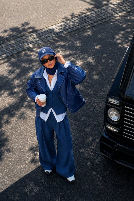 A street style photo of a person wearing an oversized denim jacket, vest, and pants, along with a blue cap and sunglasses, standing next to a black vehicle.