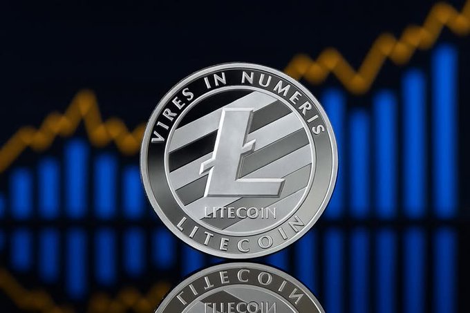 A silver Litecoin (LTC) physical coin in close-up showing the 'VIRES IN NUMERIS' Latin inscription, set against a dark background with a blue and gold stock market chart.