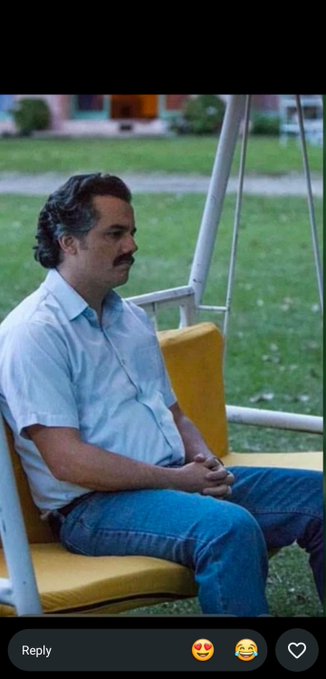 A man with a mustache wearing a light blue shirt and jeans, sitting on a yellow swing bench with a serious expression; the image has a 'Reply' comment section at the bottom.