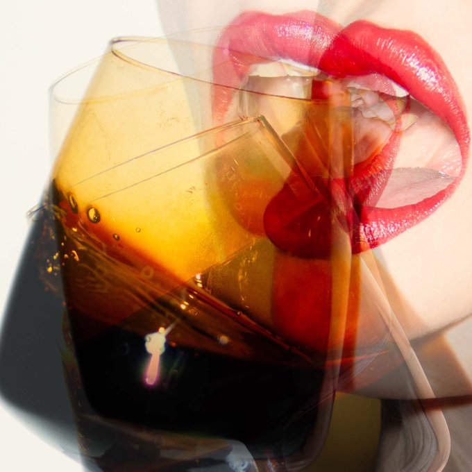 The image features a close-up view of a woman's face with her lips open wide, revealing her red lipstick. The background of the photo is blurred, but there are two wine glasses present - one filled with a dark liquid that appears to be beer or soda, and another glass containing water. The combination of these elements creates an interesting visual effect, as if the woman's lips were made of glass.