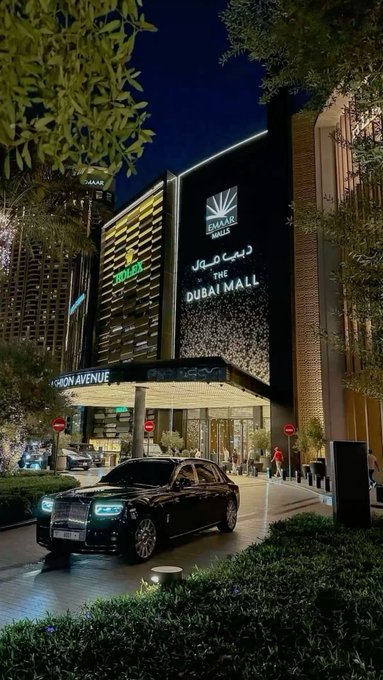 A sleek black Rolls-Royce Cullinan is parked in front of "THE DUBAI MALL" entrance at night, with the Emaar logo and "Dress Up" signage visible, alongside "ADDRESS" and "FASHION AVENUE" text, fitting the "GN" (Gulf News) post capturing Dubai's luxury lifestyle.