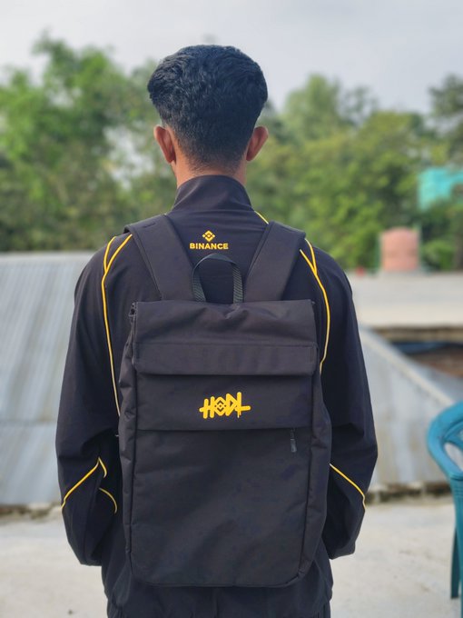 A young man stands with his back to the camera, wearing a black Binance-branded jacket and a matching black backpack featuring yellow "HSKL" lettering and the Binance logo, showcasing his first gift received from Binance.