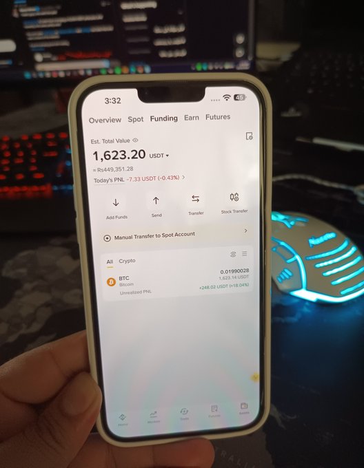 A hand holding a smartphone displaying a crypto portfolio overview screen with an estimated total value of 1,623.20 USDT, showing BTC holdings of 0.01990028, set against a background with a backlit keyboard and computer monitor.