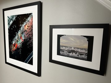 Two framed artworks hang on a gray wall, displayed side by side with black frames and white matting. The left piece features a vibrant, abstract composition with colorful drips and splatters against a dark background, while the right piece showcases a more subdued landscape scene with a muted, atmospheric quality—likely showcasing Dave Krugman's photographic work from the "Drip Drop" collection featured on FWA.