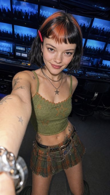 A young woman with orange-streaked black hair, star necklaces, and visible tattoos takes a selfie in what appears to be a control room filled with video monitors showing silhouettes of a crowd. The caption references her work in the "edit suite" processing footage for her audience, matching the behind-the-scenes vibe of someone handling rapid post-event content after her token sale attracted thousands of holders.