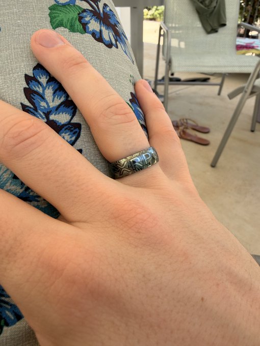 A hand displays a metallic ring engraved with "XRP" worn on the middle finger, shown against a floral-patterned cushion. The image visually demonstrates the durability and longevity of the XRP-branded ring after two years of daily use.