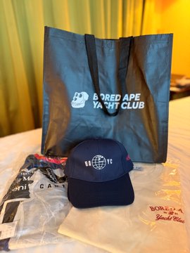 A Bored Ape Yacht Club branded shopping bag with a matching navy blue cap, displayed on a bed with packaging materials.