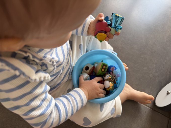 A toddler in a blue and white striped shirt holds a toy penguin while sitting with a blue bucket full of colorful toys. The image captures a sweet moment of childhood play, perfectly complementing the heartfelt caption about the child's cherished possession—the bucket of Pudgy Penguins that always brings a smile.