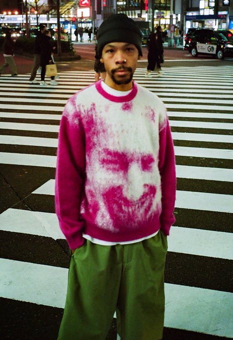 A man wearing a black beanie, olive green pants, and a pink and white sweater featuring a large skull-like print stands in the middle of a crosswalk on a city street at night. A taxi marked "GO" is visible in the background.