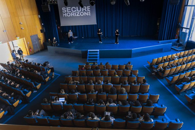 An aerial view of a 'SECURE HORIZONS' event in an auditorium with an audience seated in blue chairs watching presenters on a stage.