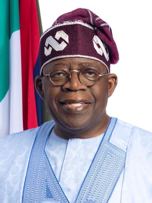An official portrait of Nigerian President Bola Ahmed Tinubu, wearing a traditional cap and light blue agbada attire, standing in front of the Nigerian flag.