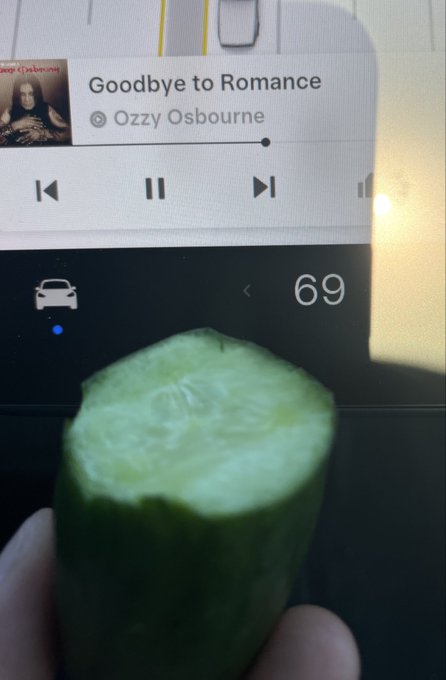 A hand holds a partially eaten cucumber in front of a Tesla dashboard display showing a temperature of 69 degrees, with music playing "Goodbye to Romance" by Ozzy Osbourne—matching the caption's casual scene of snacking and cruising rather than watching Bitcoin's price.