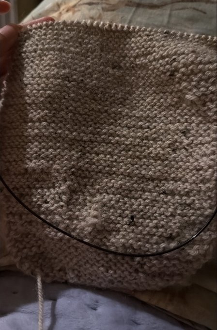 A close-up of hands working on a beige knitted fabric with a circular knitting needle, showing the textured stitch pattern.