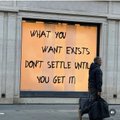 A grainy photograph shows a person in a dark jacket standing next to a brightly lit yellow sign with black handwritten text that reads 'WHAT YOU WANT EXISTS DON'T SETTLE WITH YOU GET IT'.