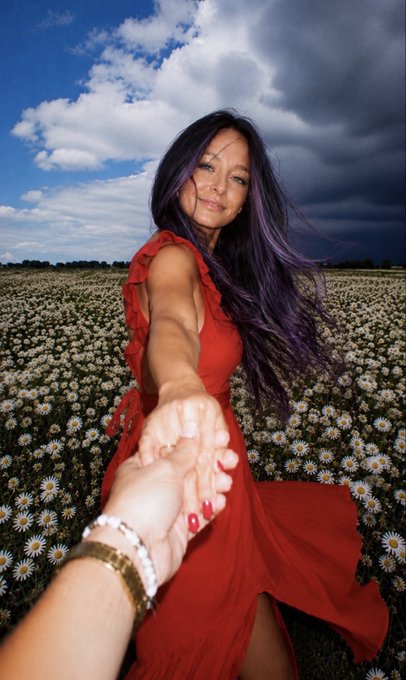 A woman with long purple-streaked hair in a flowing red dress reaches toward the camera to hold someone's hand in a field of daisies under a partly cloudy sky.