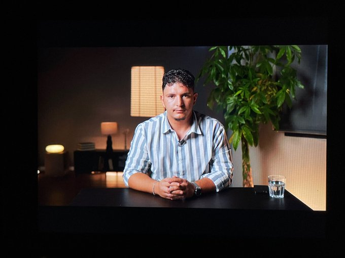 A man in a blue and white striped shirt sits at a table in a dimly lit room with a plant and lamps in the background, photographed through what appears to be a framed view.