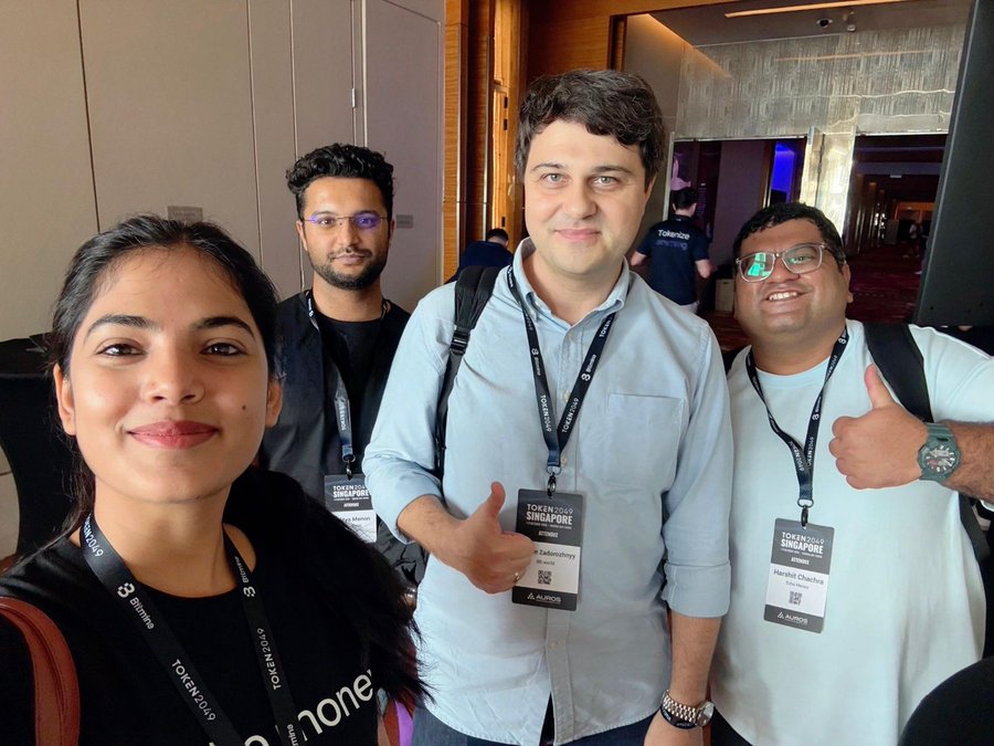 A selfie of four smiling people at the TOKEN2049 Singapore conference, wearing branded lanyards including attendees named Harshil Chudnak and what appears to be a Bitfinex attendee.