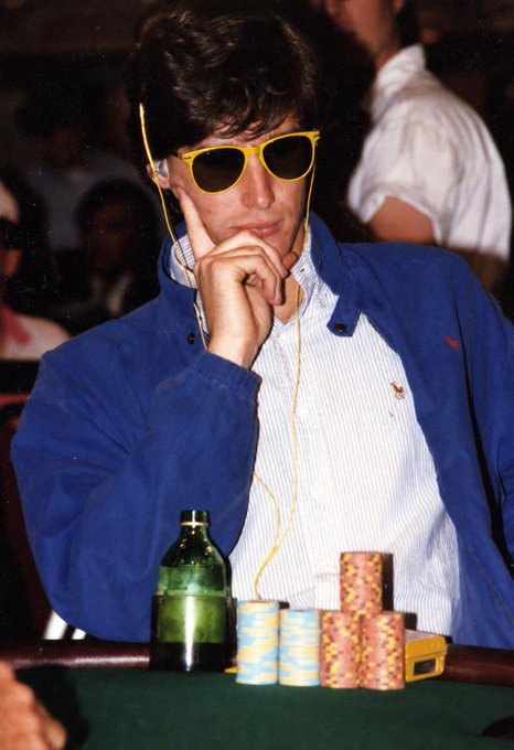A young man wearing yellow sunglasses and a blue cardigan sits at a poker table with stacks of colorful casino chips and a green glass bottle in front of him, with a yellow earphone cord visible.