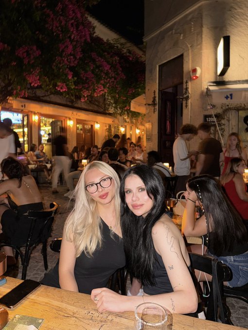 Two women pose together at a lively outdoor restaurant or café at night, with string lights, bougainvillea, and other patrons visible in the background; the number "1201" and "+1" can be seen on signs nearby, while the woman on the right displays a "MONEY" tattoo on her arm. The nighttime social scene complements the post's bullish "GM" (good morning) sentiment about holding $SOL rather than selling.