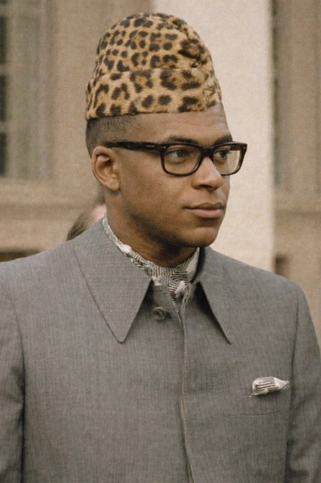 A historical photograph of a young man wearing a leopard-print kufi cap, black-rimmed glasses, and a gray suit jacket.