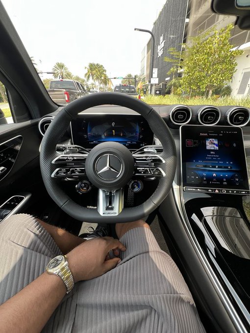 Interior view from the passenger seat of a Mercedes-AMG vehicle showing the steering wheel, digital dashboard, infotainment screen playing music, and a person's arm wearing a gold and silver watch resting on their leg.