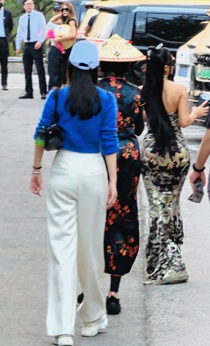Three women walk down a street; one wears a blue sweater, white pants and a light blue cap, another wears a black floral dress and a conical hat, and the third wears an ornate patterned dress.