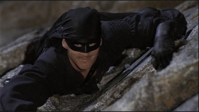 A masked man in a black costume and hat climbing up a stone wall, with a small mustache visible beneath his mask.